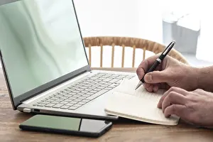 Man Working From Home Impressions: man taking notes on paper while using laptop computer